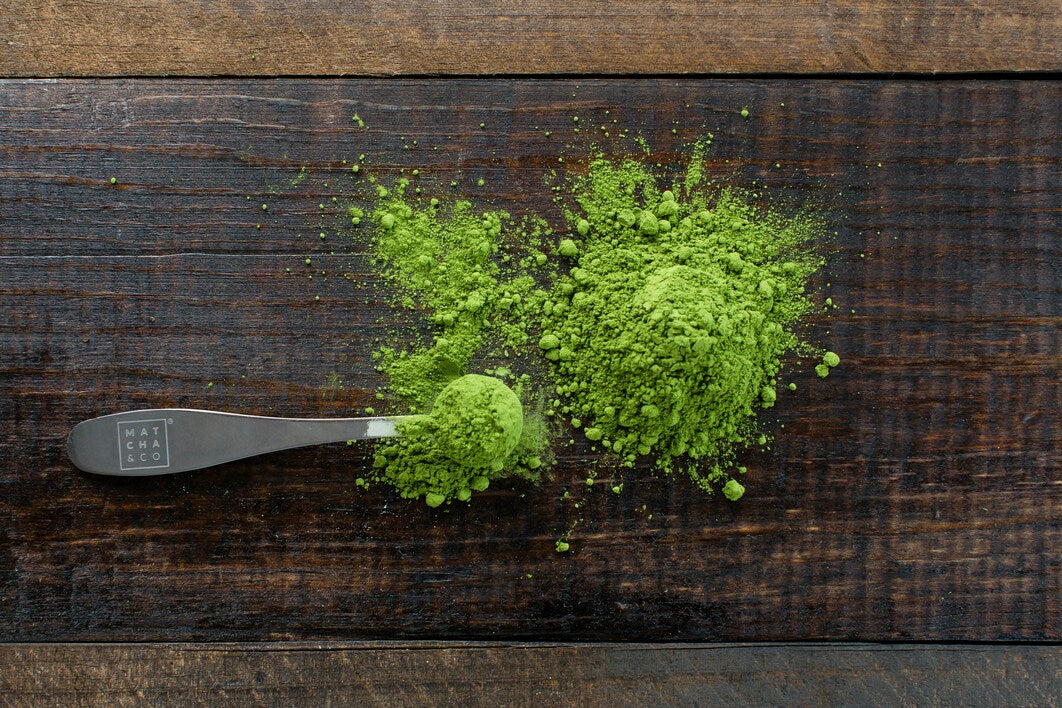 Measuring Spoon Full of Green Kratom Powder Next to Pile of Green Kratom Powder on Wooden Table