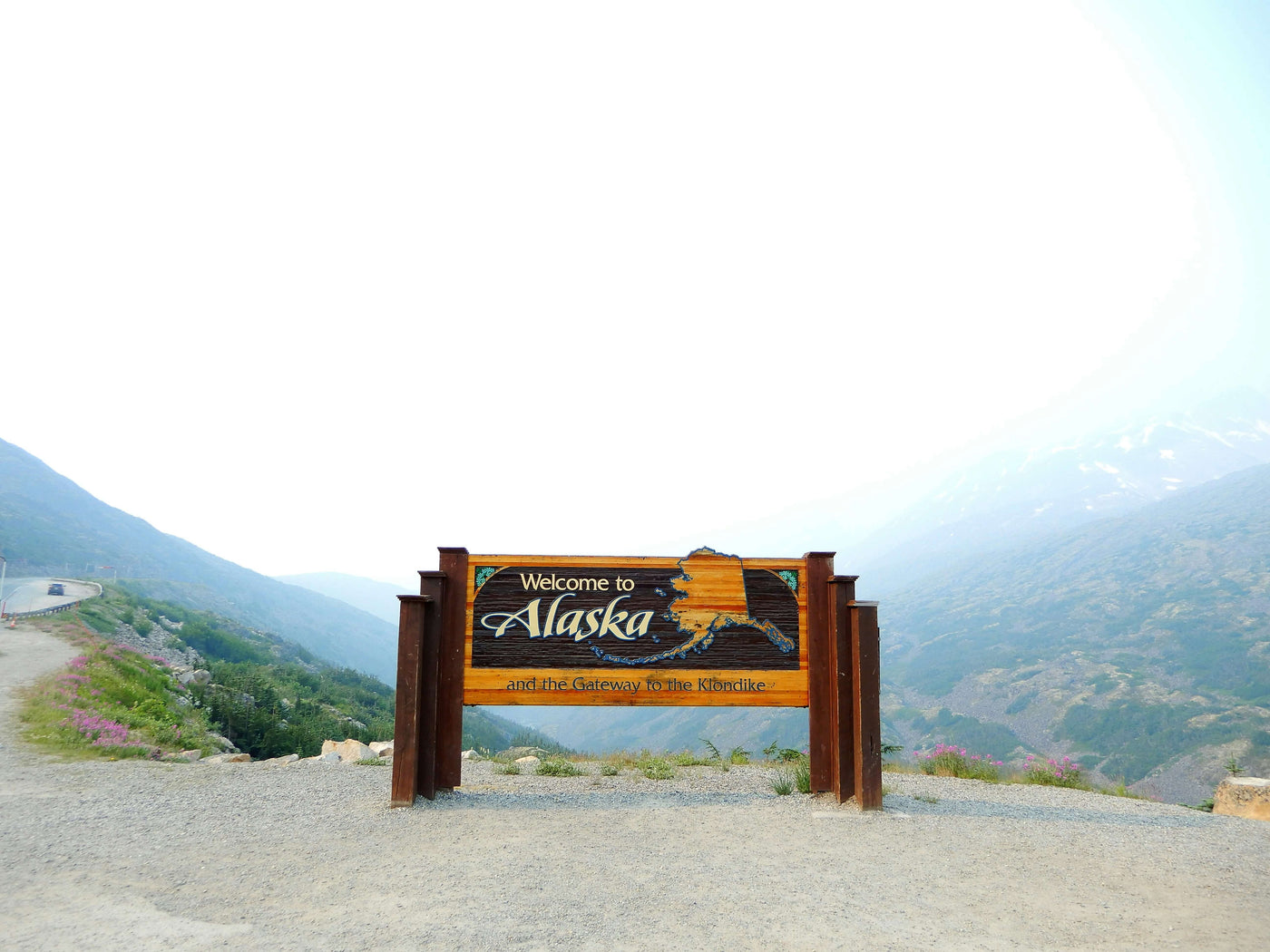 A “Welcome to Alaska” sign is prominently displayed on an overlook with a mountain backdrop